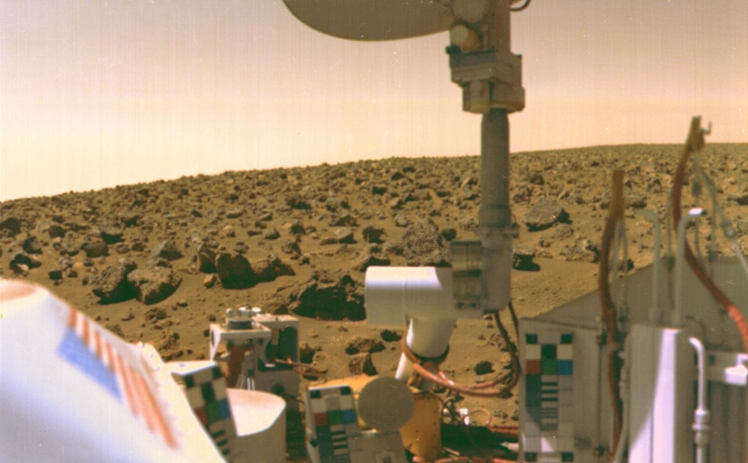 Rover’s-eye view of an orange-brown, barren landscape covered in boulders of many sizes. In the near foreground, part of the rover is captured in its own camera’s field of view.
