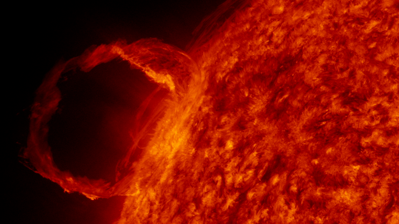 A portion of the red Sun, with a loop of solar material cascading against the dark expanse of space.